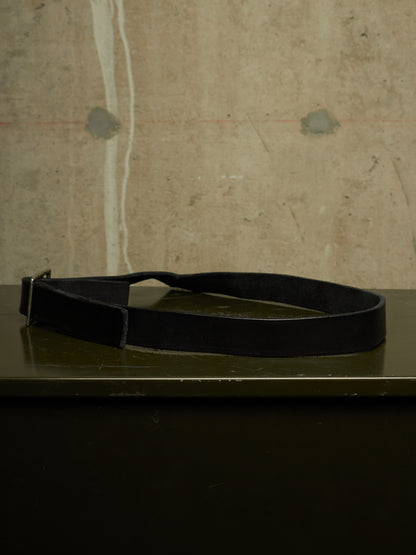 A black leather belt with a silver buckle, resting on a dark green surface against a textured concrete wall.
