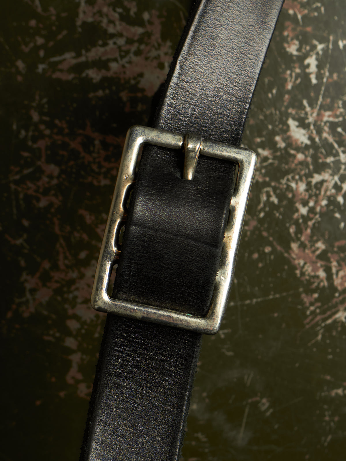 A close-up of a black leather belt with a rectangular, antique silver-tone buckle, set against a dark, textured background.