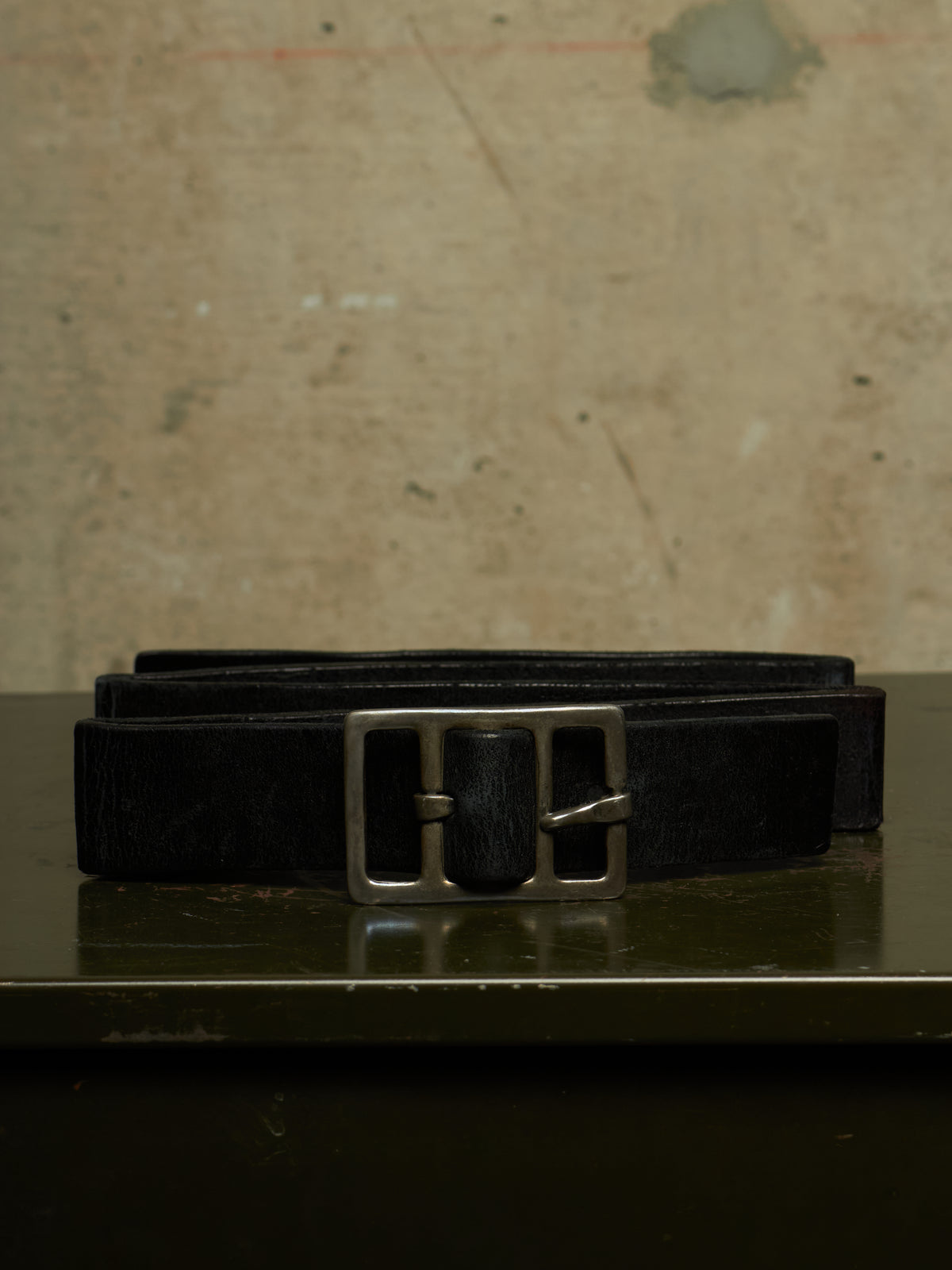 A black textured leather belt with a silver-toned metal buckle, coiled on a dark reflective surface against a textured beige wall background.