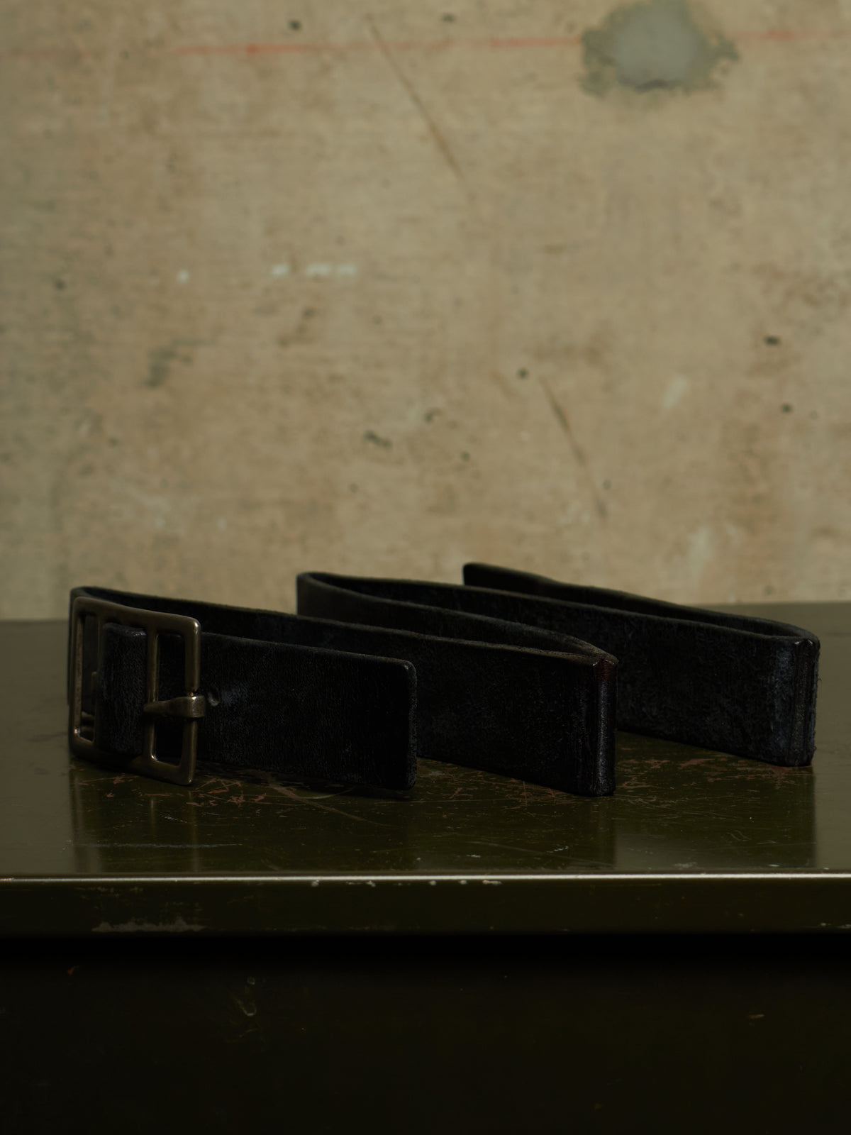 A black leather belt with a silver-toned metal buckle, resting on a dark reflective surface against a textured beige wall.