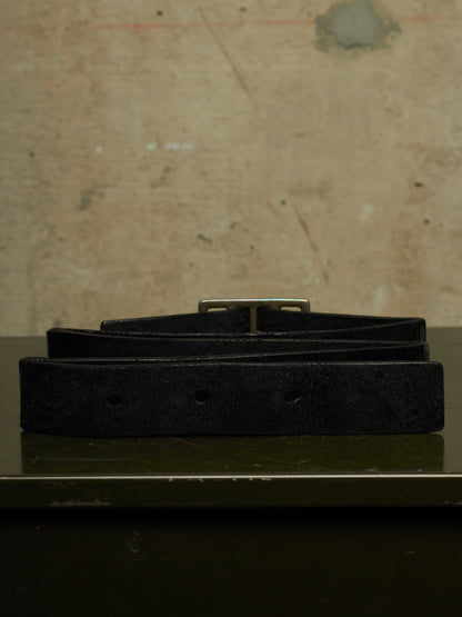 A black suede belt with a silver-tone metal buckle, neatly coiled and displayed on a dark reflective surface against a textured, warm beige background.