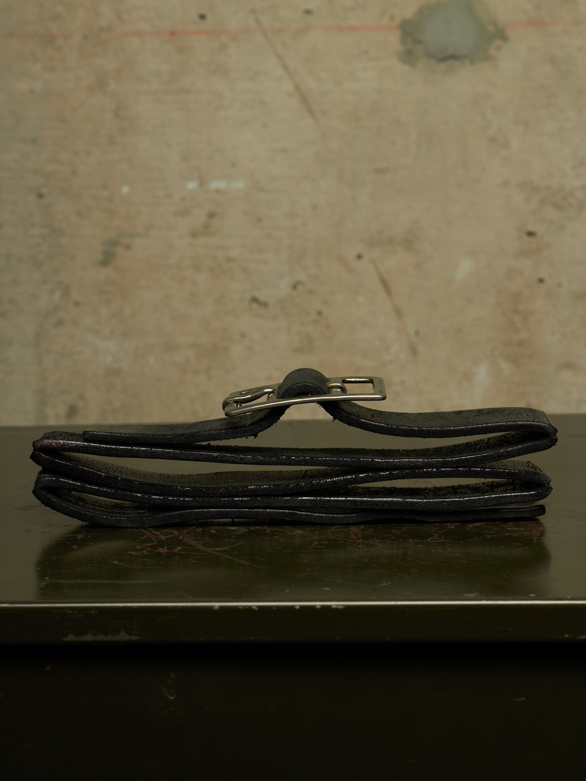 A black leather belt folded on a dark reflective surface against a textured beige wall, featuring a silver metal buckle.