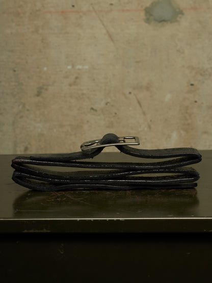 A black leather belt folded on a dark reflective surface against a textured beige wall, featuring a silver metal buckle.