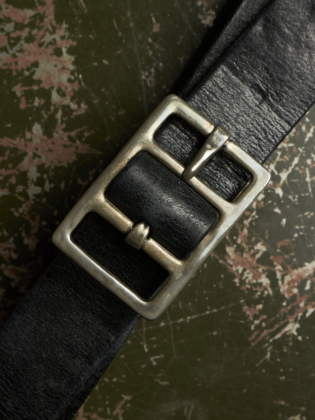 A close-up of a black leather belt with a large, rustic silver-toned metal buckle, laid diagonally across a textured, distressed green and brown background.