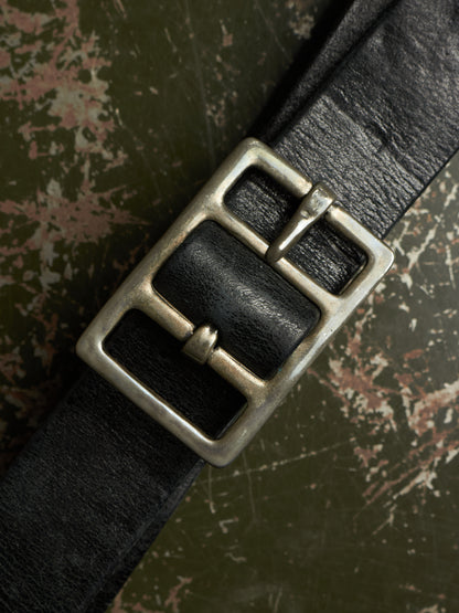 A close-up of a black leather belt with a large, rustic silver-toned metal buckle, laid diagonally across a textured, distressed green and brown background.