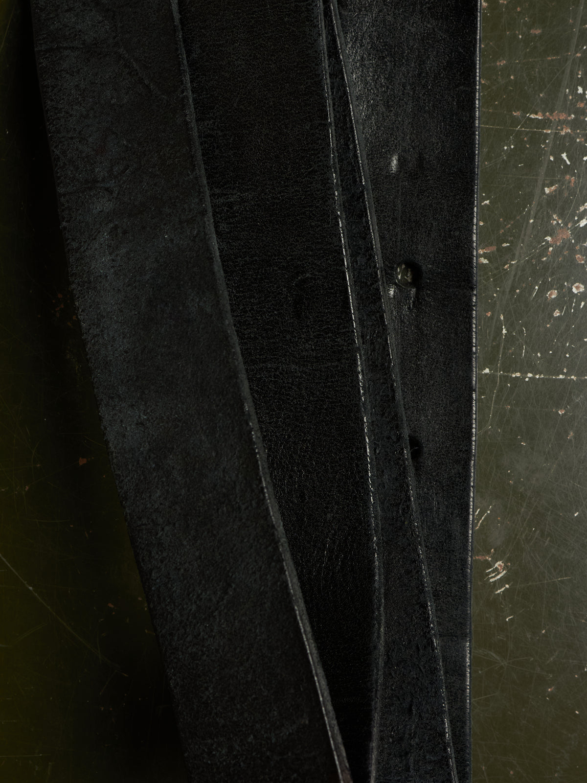 A close-up of three dark, textured leather strips or straps laid vertically against a dark, scratched metal surface.