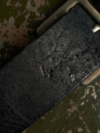 A close-up of a dark textured leather belt with a distressed, weathered finish and a silver-toned metal buckle, set against a distressed olive green background.