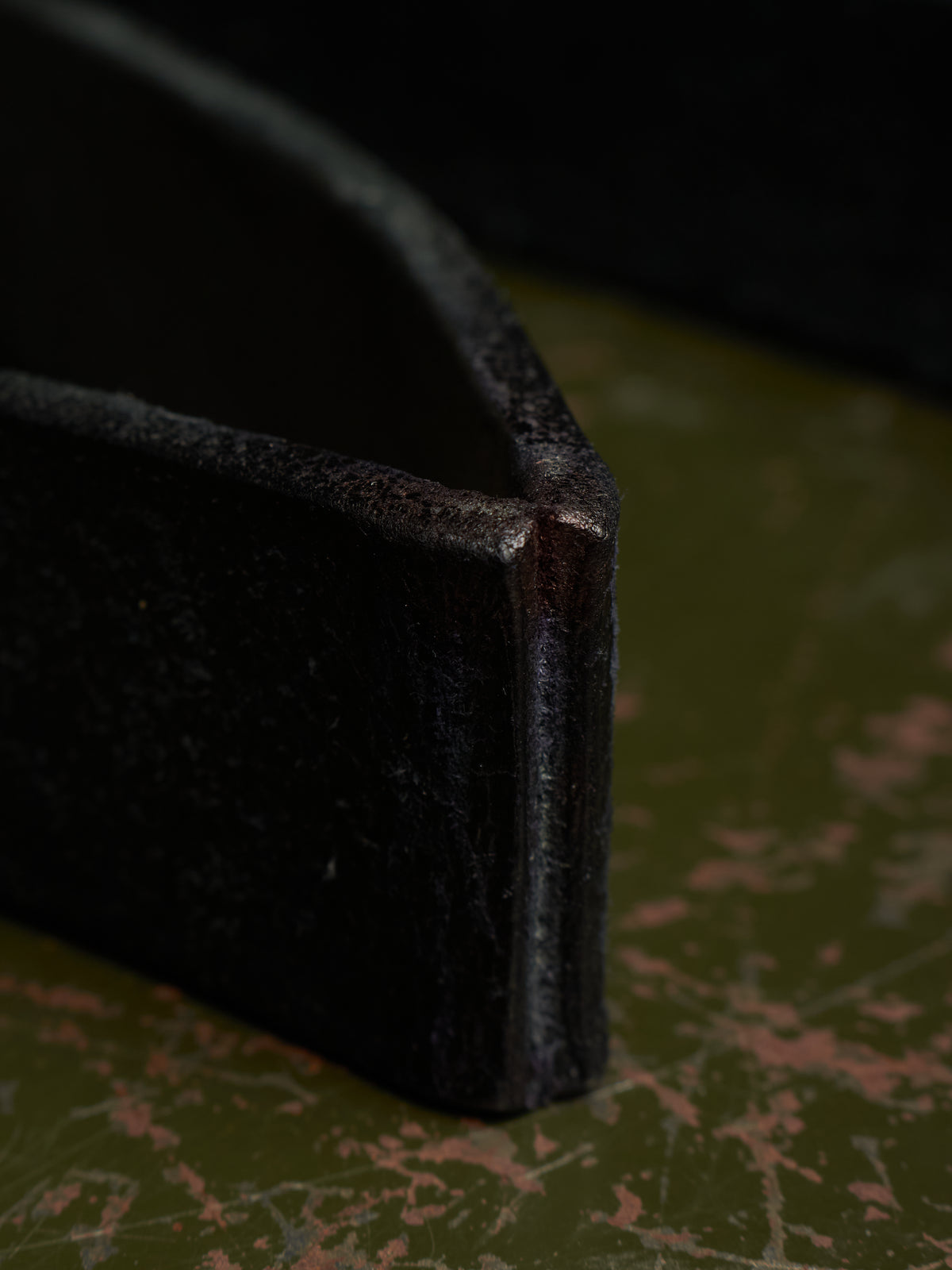 Close-up of a dark, rustic metal tray corner showing its textured, weathered surface and a sharp edge, resting on a distressed olive green surface.