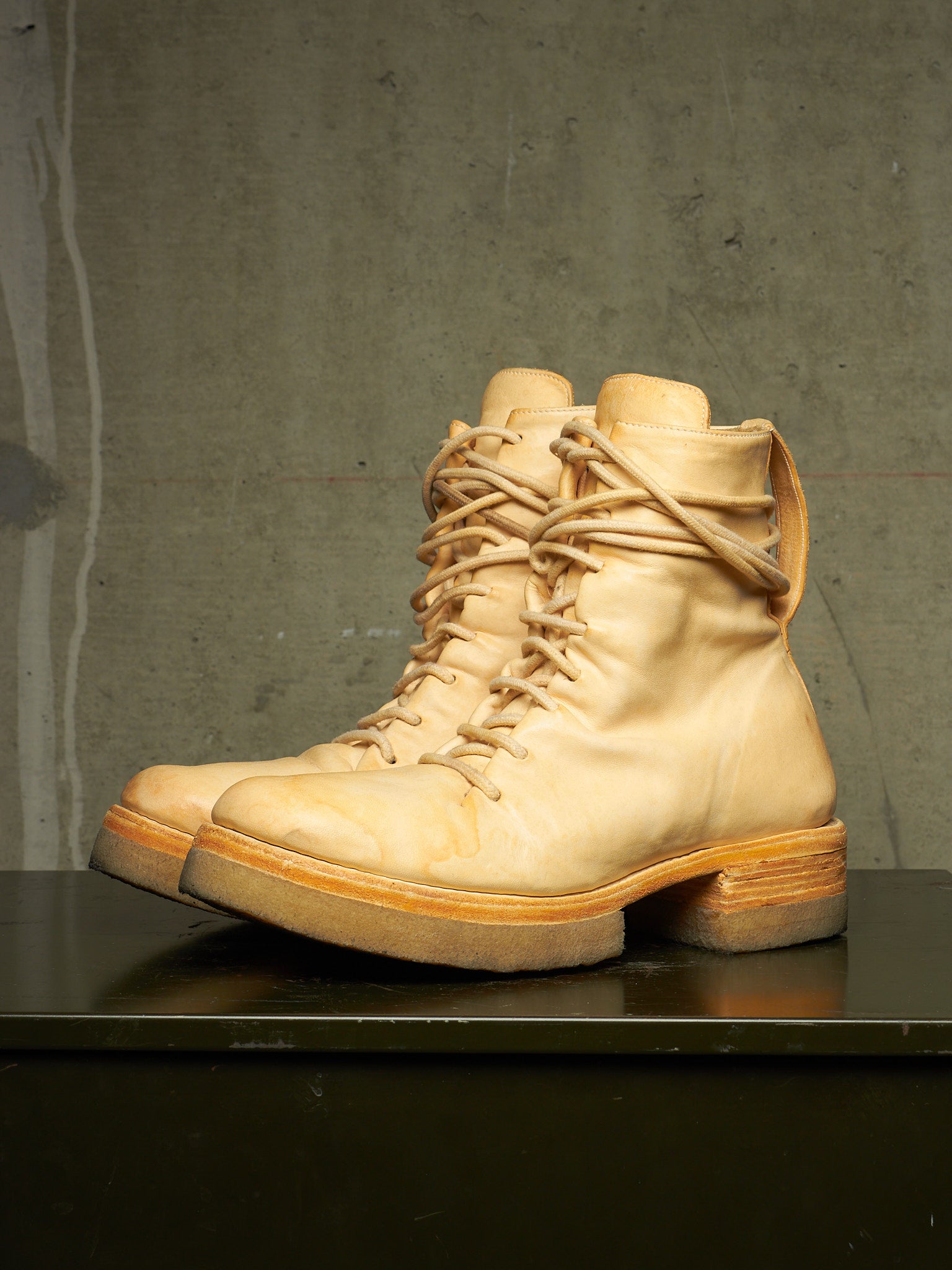 A pair of light beige leather lace-up combat boots with chunky soles, displayed on a dark surface against a textured gray concrete wall.