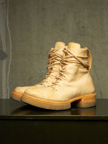 A pair of light beige leather lace-up combat boots with chunky soles, displayed on a dark surface against a textured gray concrete wall.