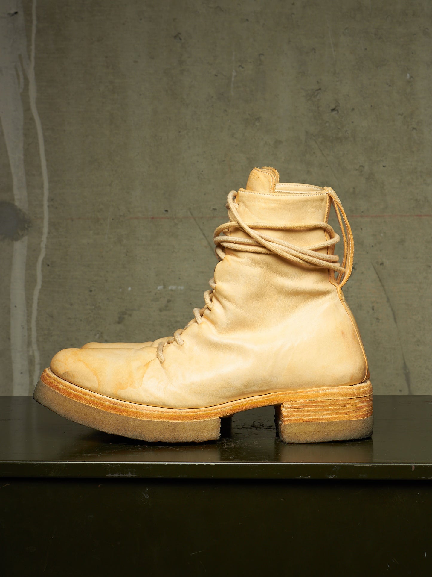 A single light beige leather lace-up boot with a stacked wooden block heel, displayed in profile on a dark surface against a textured gray concrete wall.