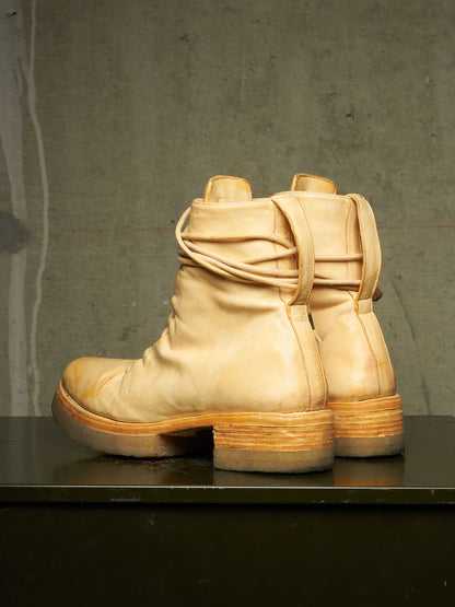 A pair of light tan leather ankle boots with draped straps and stacked wood-look heels, displayed on a dark surface against a textured gray concrete wall.