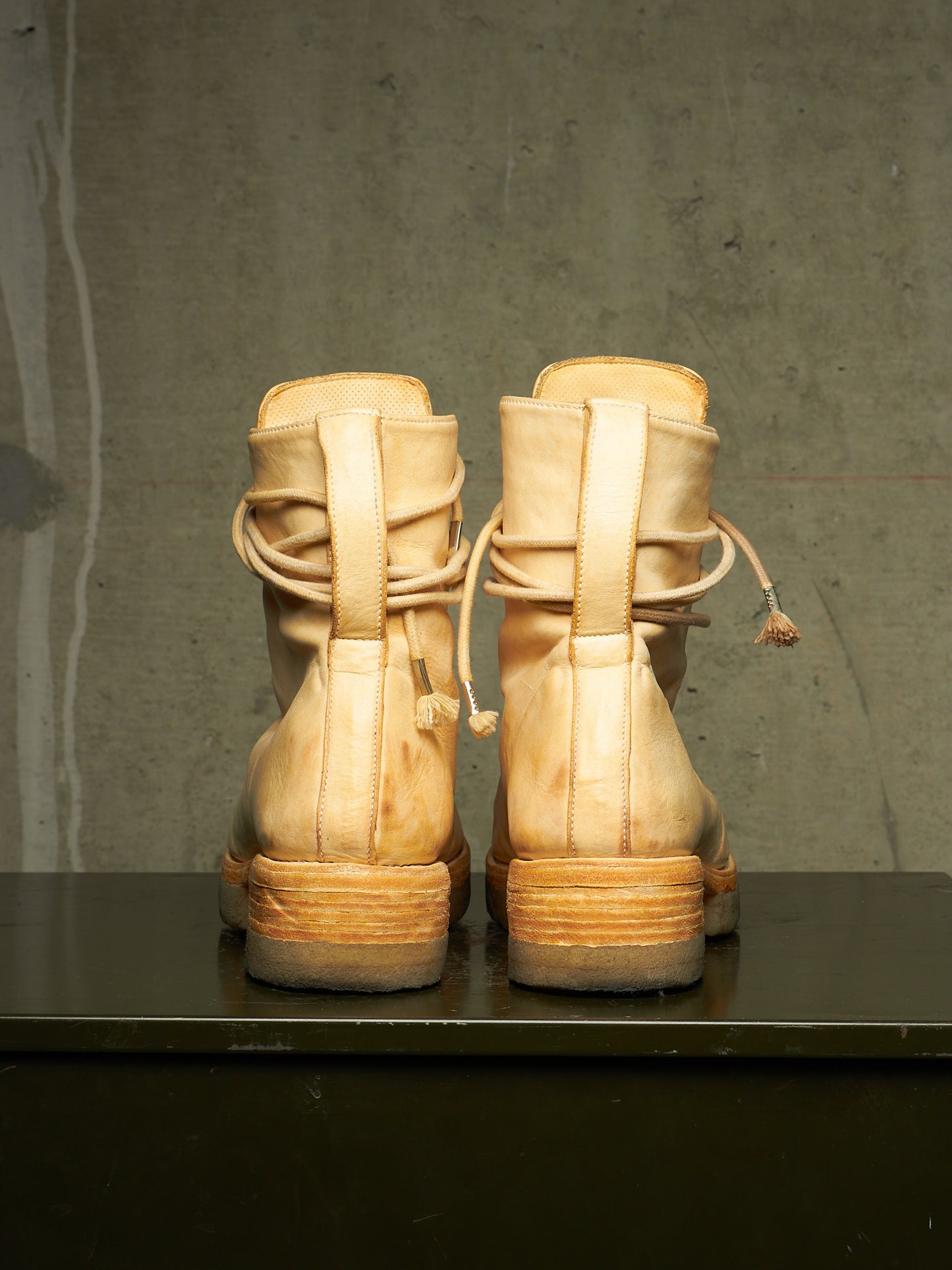 A pair of light beige leather lace-up ankle boots shown from the back, resting on a dark surface against a textured gray concrete wall.
