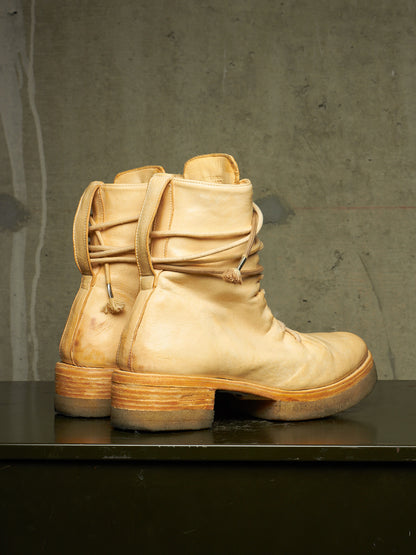 A pair of light tan leather ankle boots with stacked wooden-look heels, shown from the back on a dark reflective surface against a textured gray concrete wall.