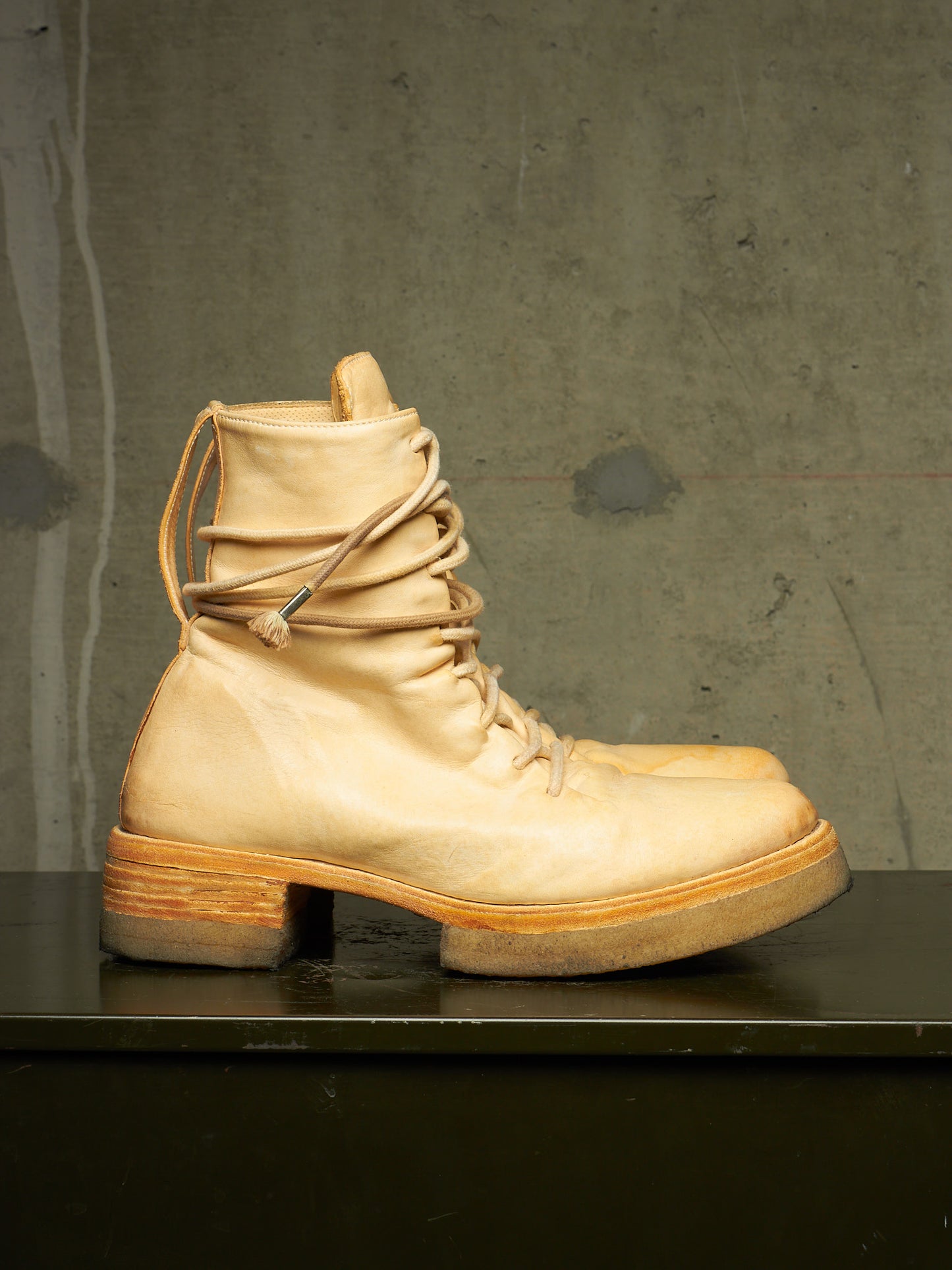 A single light beige leather lace-up boot with a chunky block heel and stacked sole, displayed on a dark surface against a textured gray concrete wall.