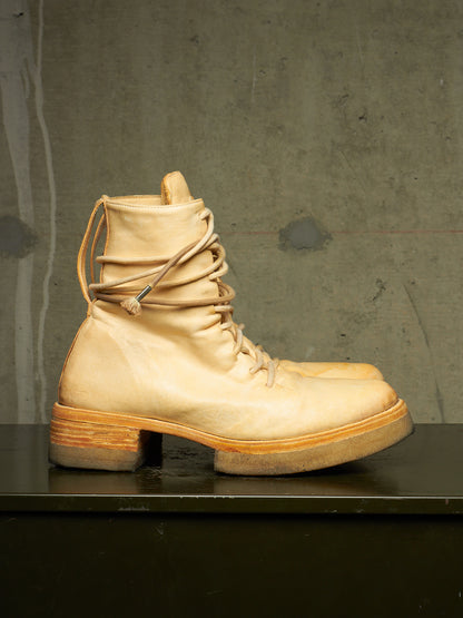 A single light beige leather lace-up boot with a chunky block heel and stacked sole, displayed on a dark surface against a textured gray concrete wall.