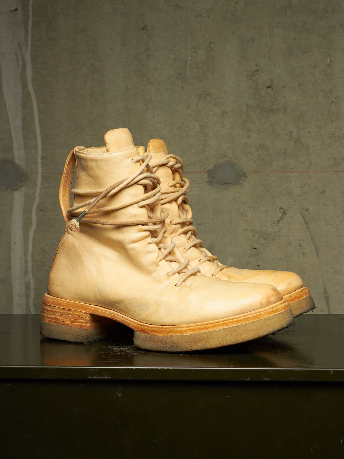 A pair of light beige leather lace-up boots with a chunky sole and stacked heel, displayed on a dark surface against a textured gray concrete wall.
