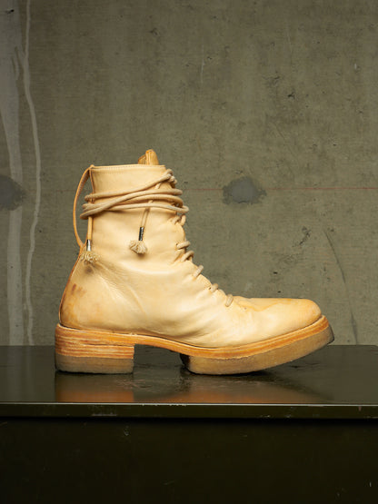 A single light beige leather lace-up boot with a chunky stacked sole and decorative hanging cord details, displayed in profile on a dark surface against a textured gray wall.