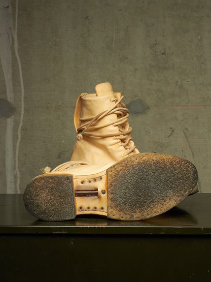A single light tan leather lace-up boot shown from the sole side, resting on a dark surface against a textured gray concrete wall.