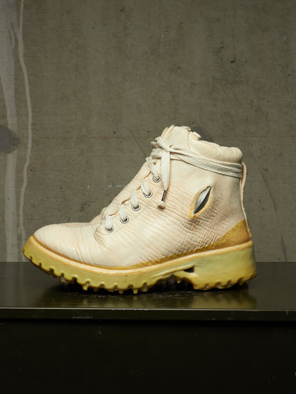 A single high-top lace-up boot in off-white textured leather with a yellowed sole and a small metallic accent on the side, displayed on a dark surface against a gray concrete wall.