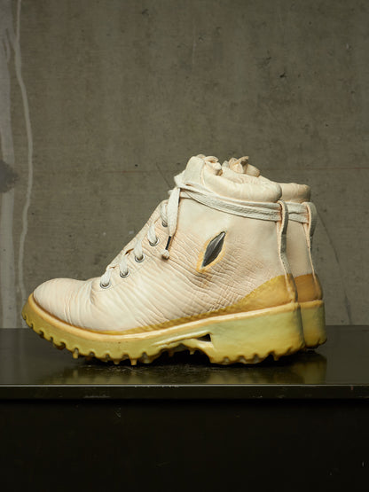 A single high-top lace-up boot in off-white leather with a yellowed sole and heel, displayed on a dark reflective surface against a textured gray concrete wall.