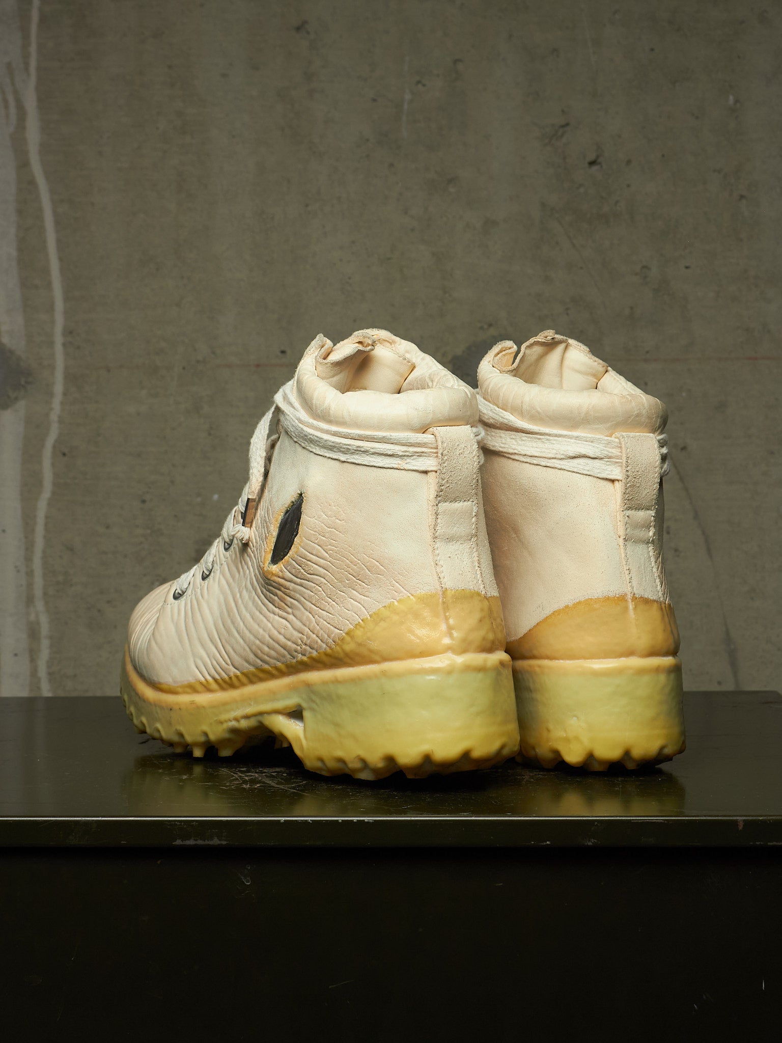 A pair of off-white leather high-top boots with yellow-tan rubber soles, shown from the back on a dark reflective surface against a textured gray concrete wall.