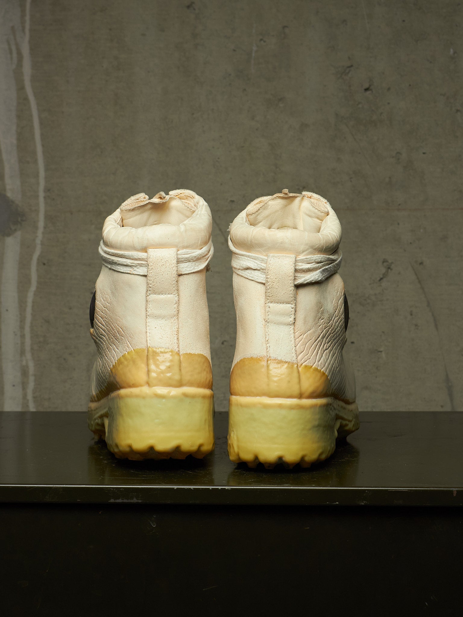 A pair of high-top sneakers shown from the back, featuring off-white leather uppers with a yellowed, aged sole and heel cup, set against a dark gray concrete wall and black surface.