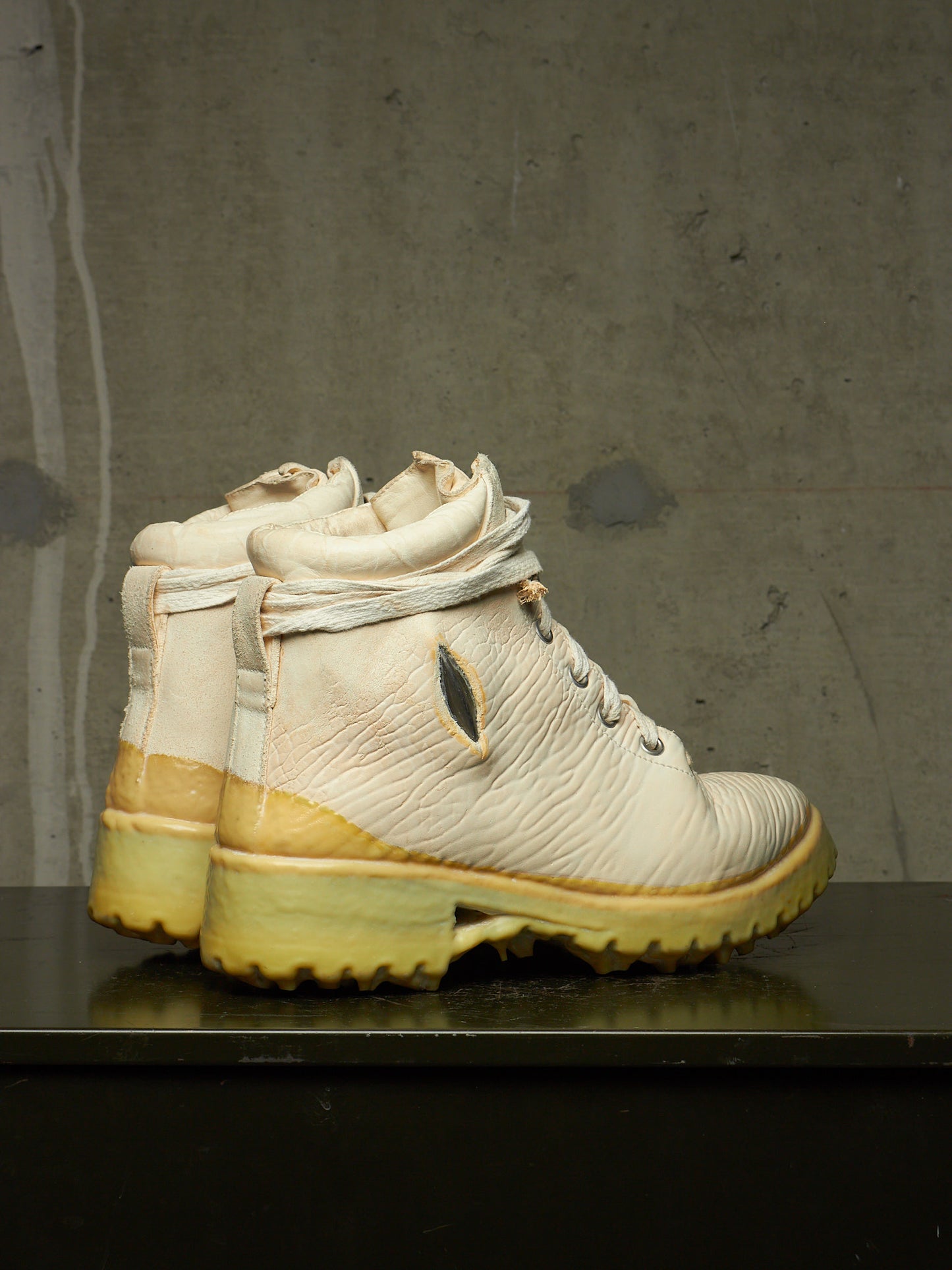 A pair of off-white leather high-top boots with yellow soles, displayed on a dark reflective surface against a textured gray concrete wall.