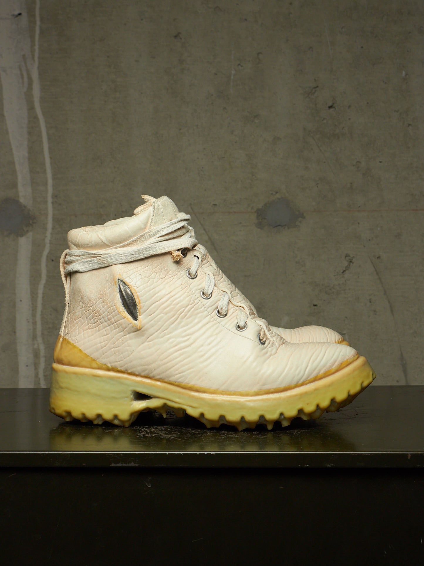 A single high-top boot in off-white textured leather with a yellow-tinted sole and heel, photographed on a dark surface against a gray concrete wall.
