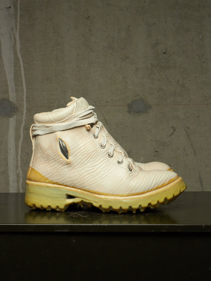 A single high-top boot in off-white textured leather with a yellow-tinted sole and heel, photographed on a dark surface against a gray concrete wall.