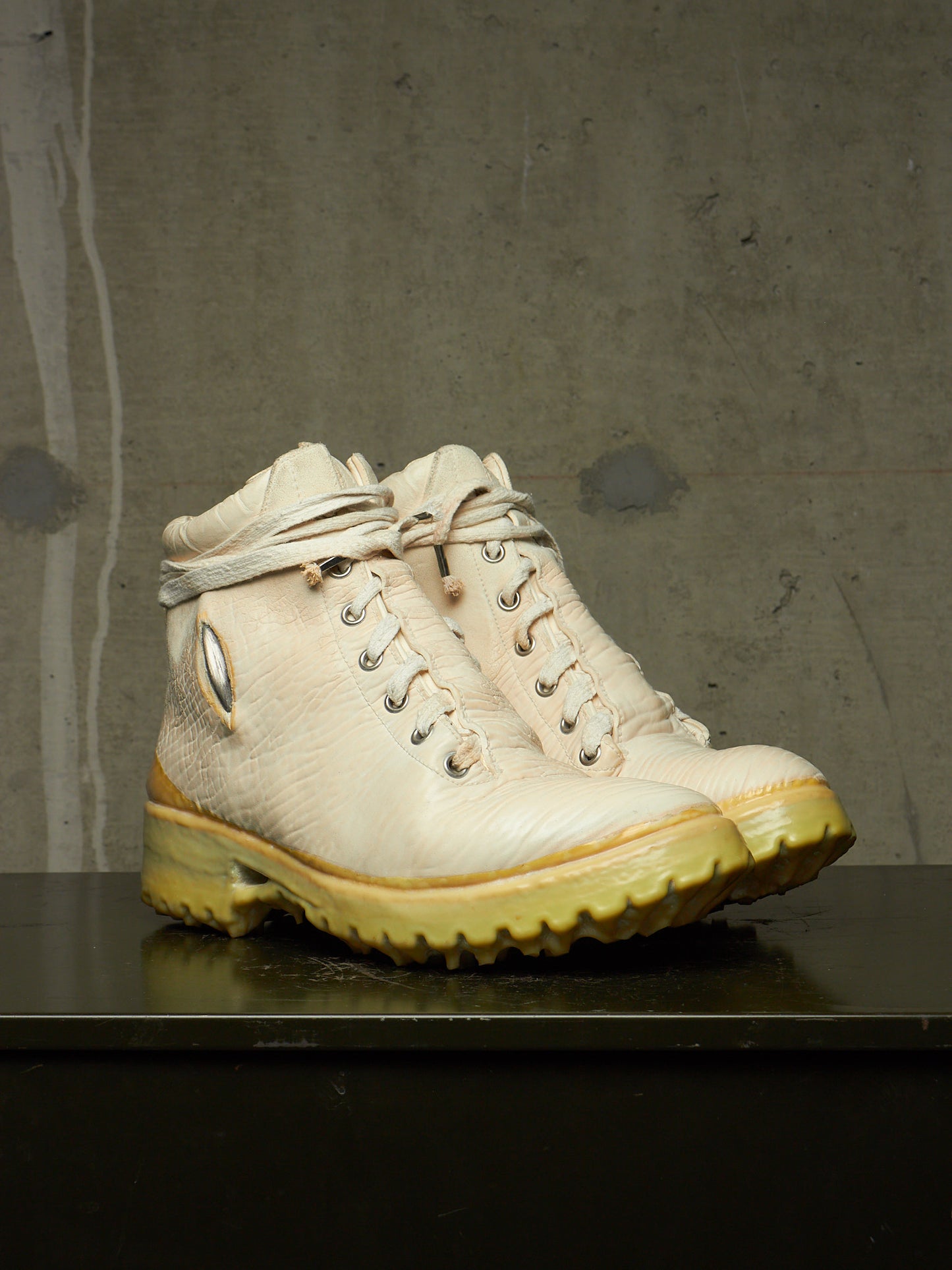 A pair of off-white leather lace-up boots with yellow soles, displayed on a dark surface against a textured gray concrete wall.