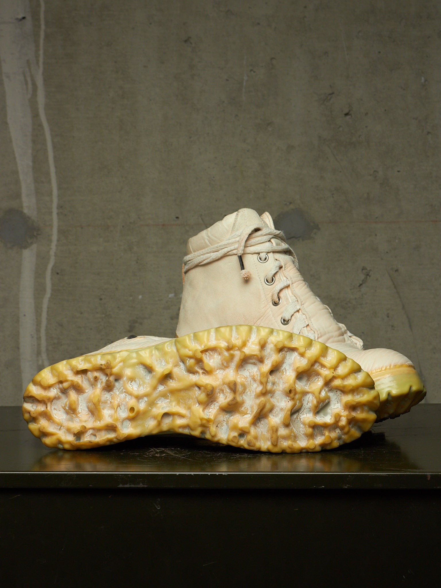 A pair of off-white lace-up boots with highly textured, yellowed tumbled soles, displayed on a dark surface against a gray concrete wall.