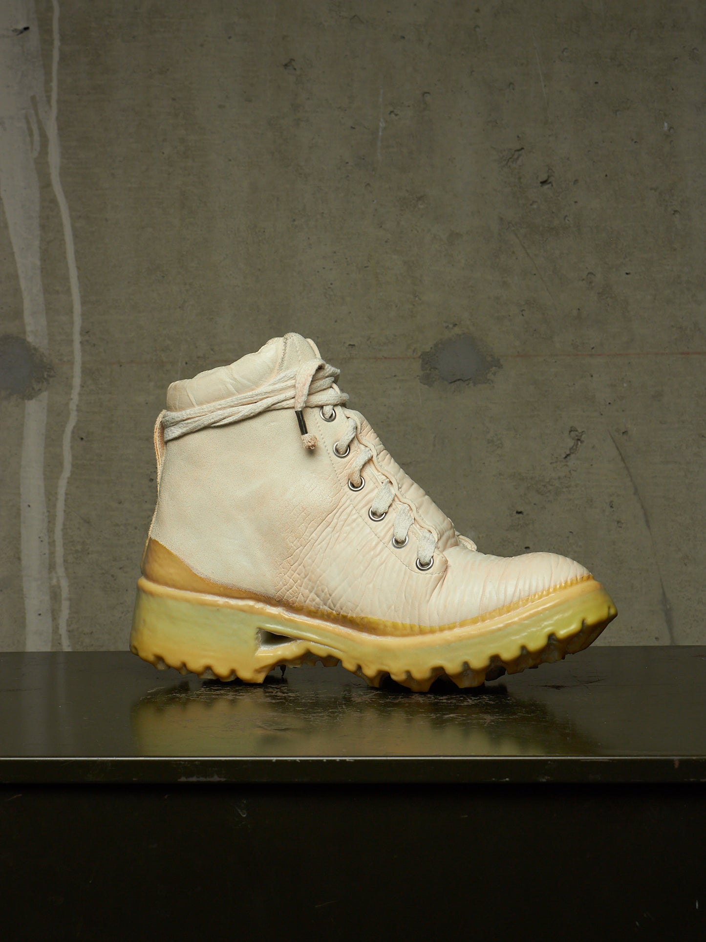 A single high-top lace-up boot in off-white leather with a thick yellow-tinted sole, resting on a dark reflective surface against a textured gray concrete wall.