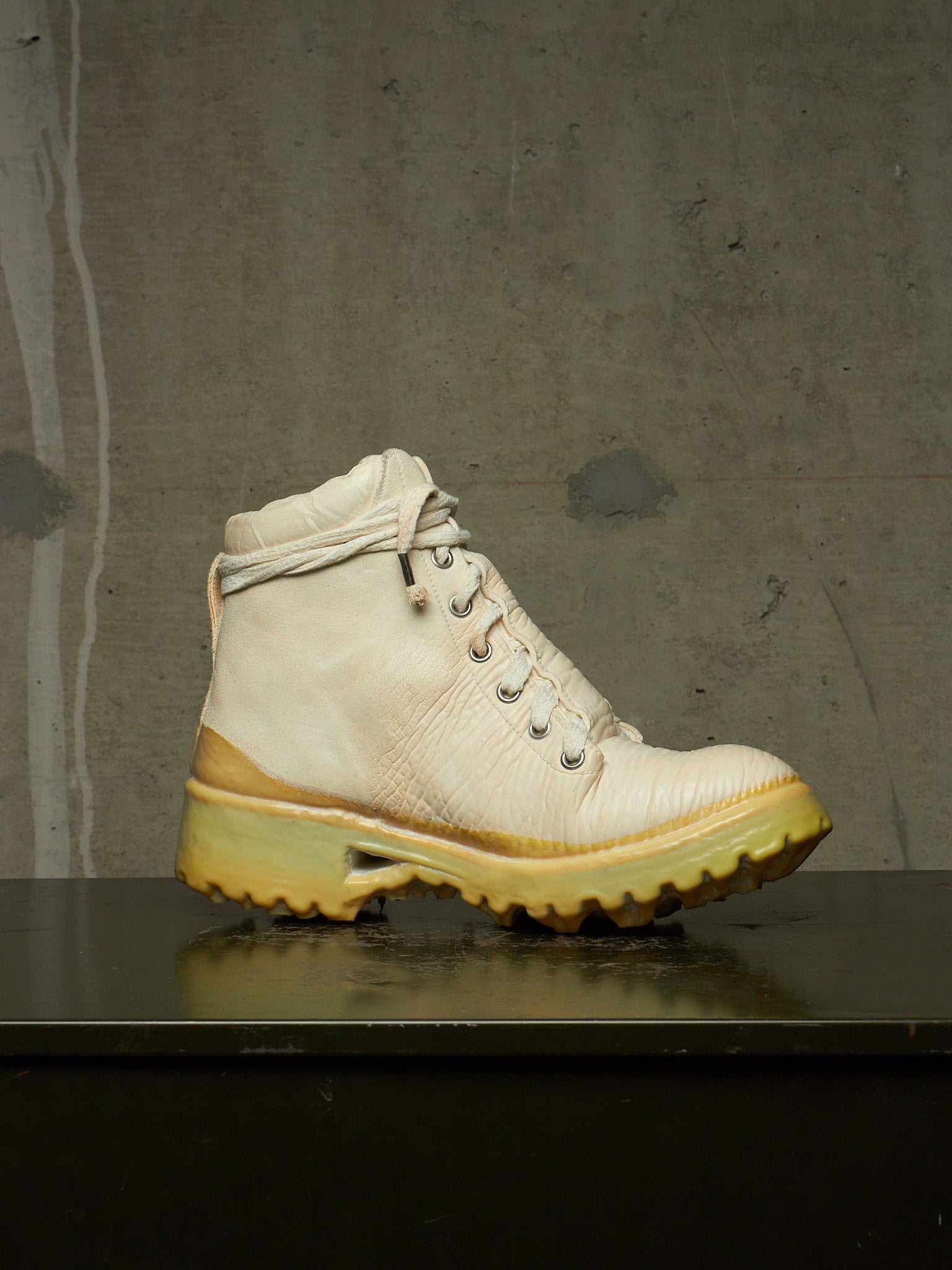 A single high-top lace-up boot in off-white leather with a thick yellow-tinted sole, resting on a dark reflective surface against a textured gray concrete wall.