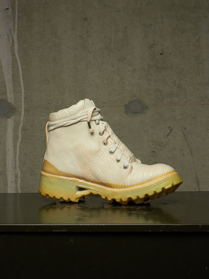 A single high-top lace-up boot in off-white leather with a thick yellow-tinted sole, resting on a dark reflective surface against a textured gray concrete wall.