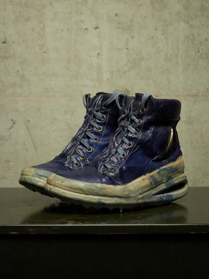 A pair of heavily distressed, vintage-style blue leather lace-up boots with thick soles, displayed on a dark surface against a textured concrete wall.