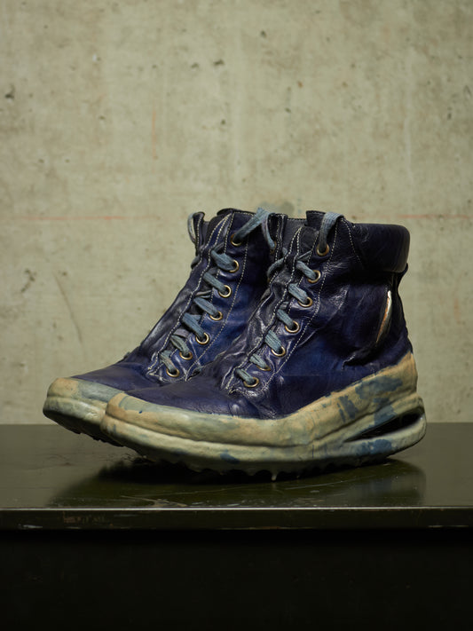A pair of heavily distressed, vintage-style blue leather lace-up boots with thick soles, displayed on a dark surface against a textured concrete wall.