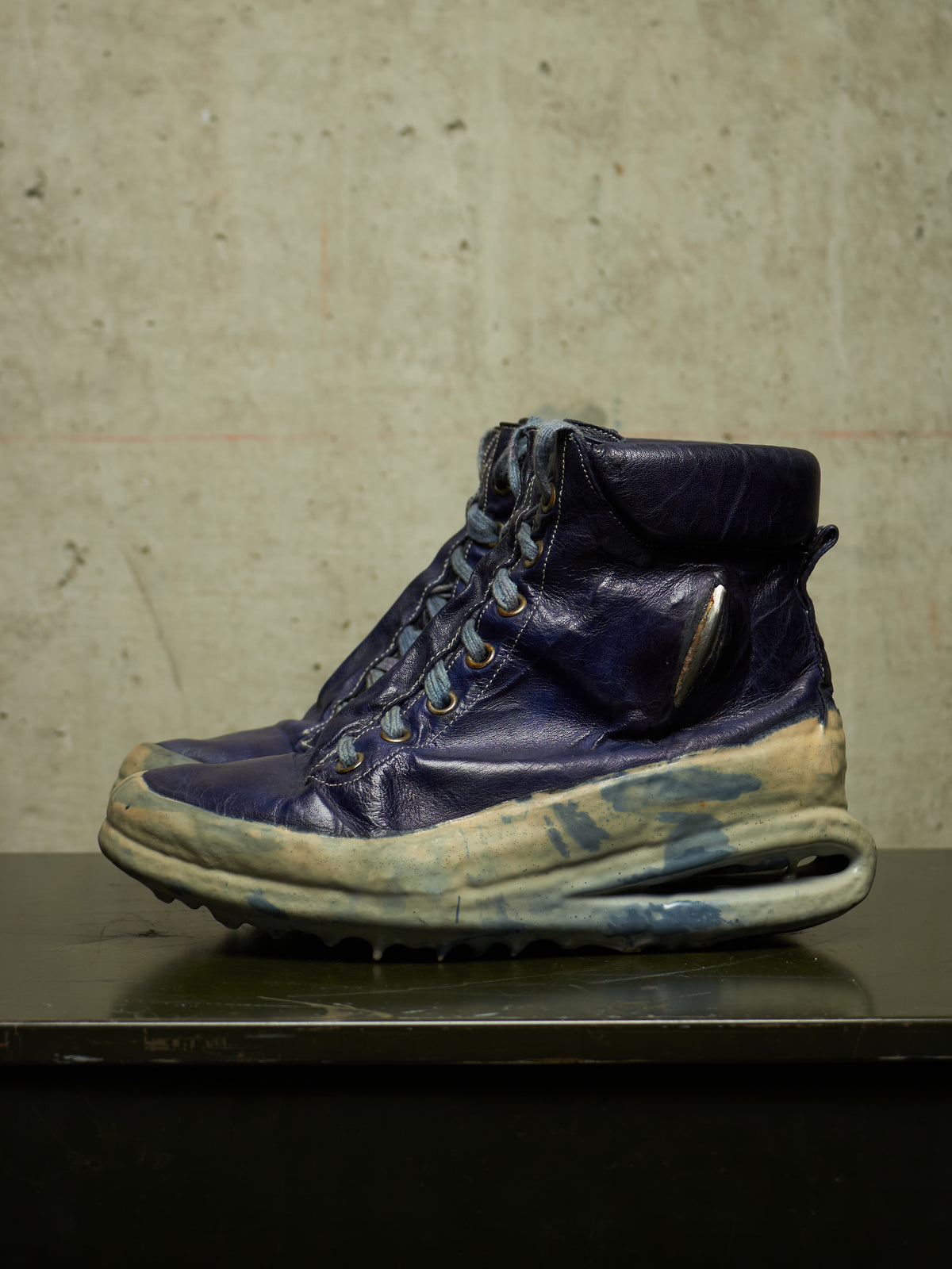 A single high-top sneaker boot in distressed navy blue leather with a chunky, sculpted sole, resting on a dark reflective surface against a textured concrete wall.