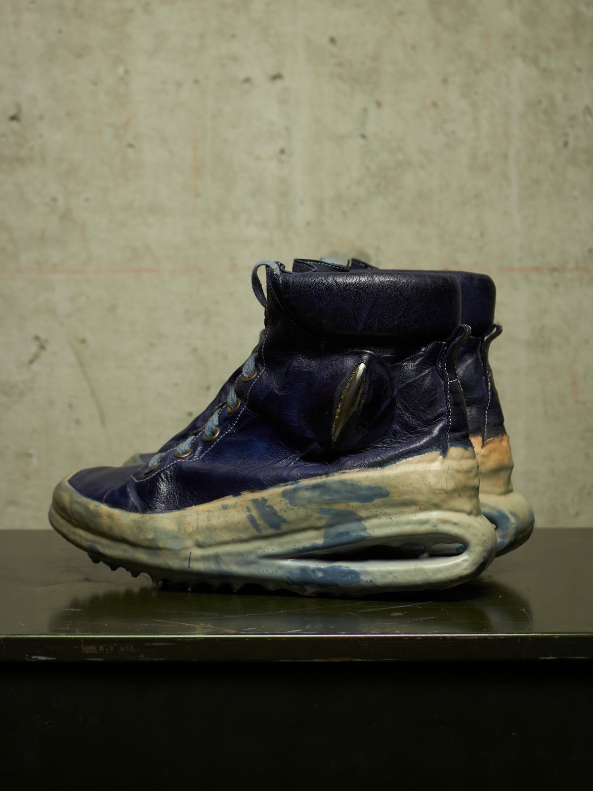 A single high-top sneaker boot with a dark blue leather upper, a thick sculptural sole in cream and blue, and a circular metallic accent on the side, displayed on a dark surface against a textured concrete wall.