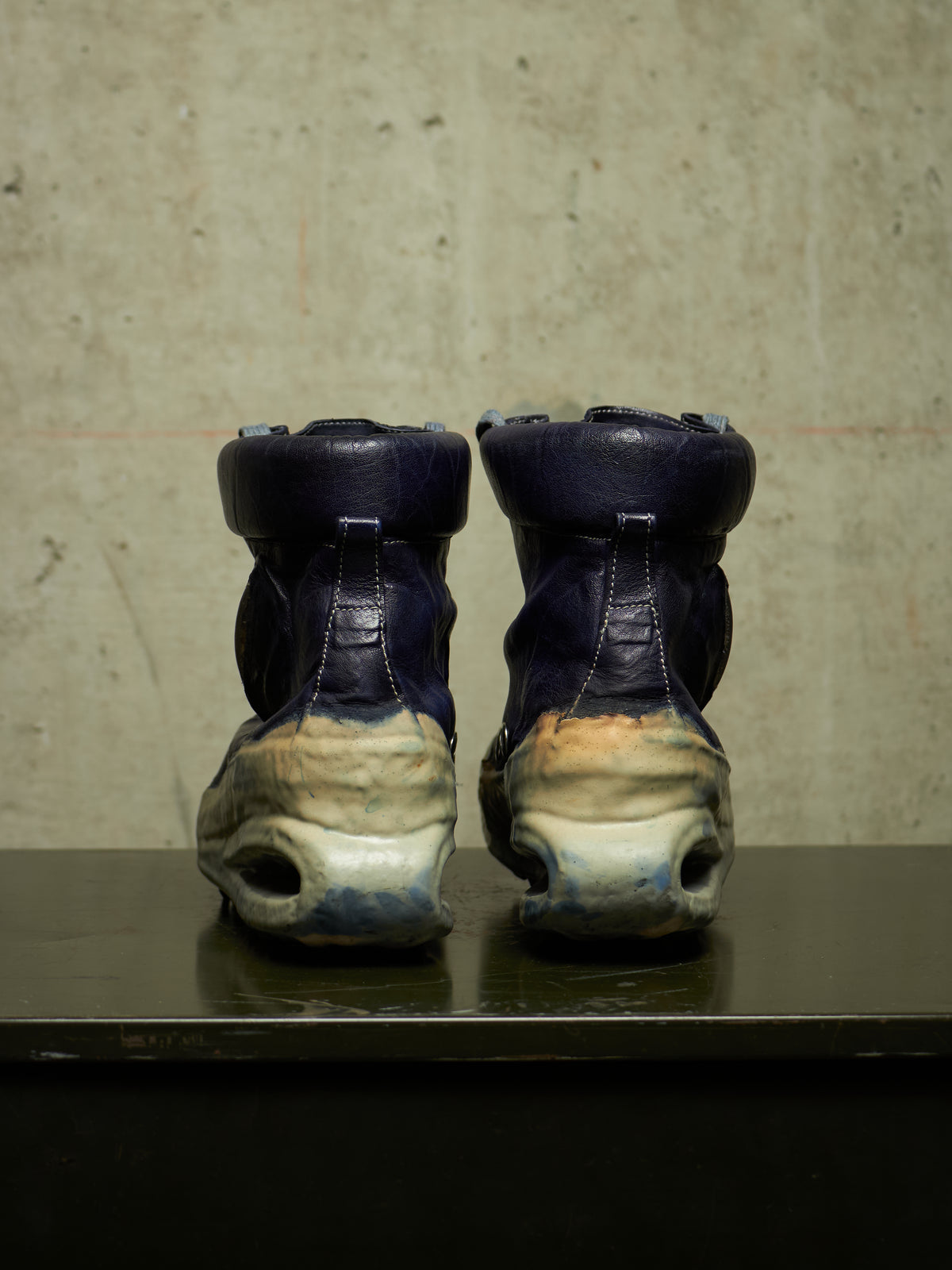 A pair of high-top sneakers shown from the back, featuring dark navy blue leather uppers and heavily distressed off-white and beige chunky soles, displayed on a dark reflective surface against a textured concrete wall.