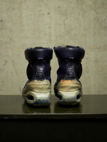 A pair of high-top sneakers shown from the back, featuring dark navy blue leather uppers and heavily distressed off-white and beige chunky soles, displayed on a dark reflective surface against a textured concrete wall.