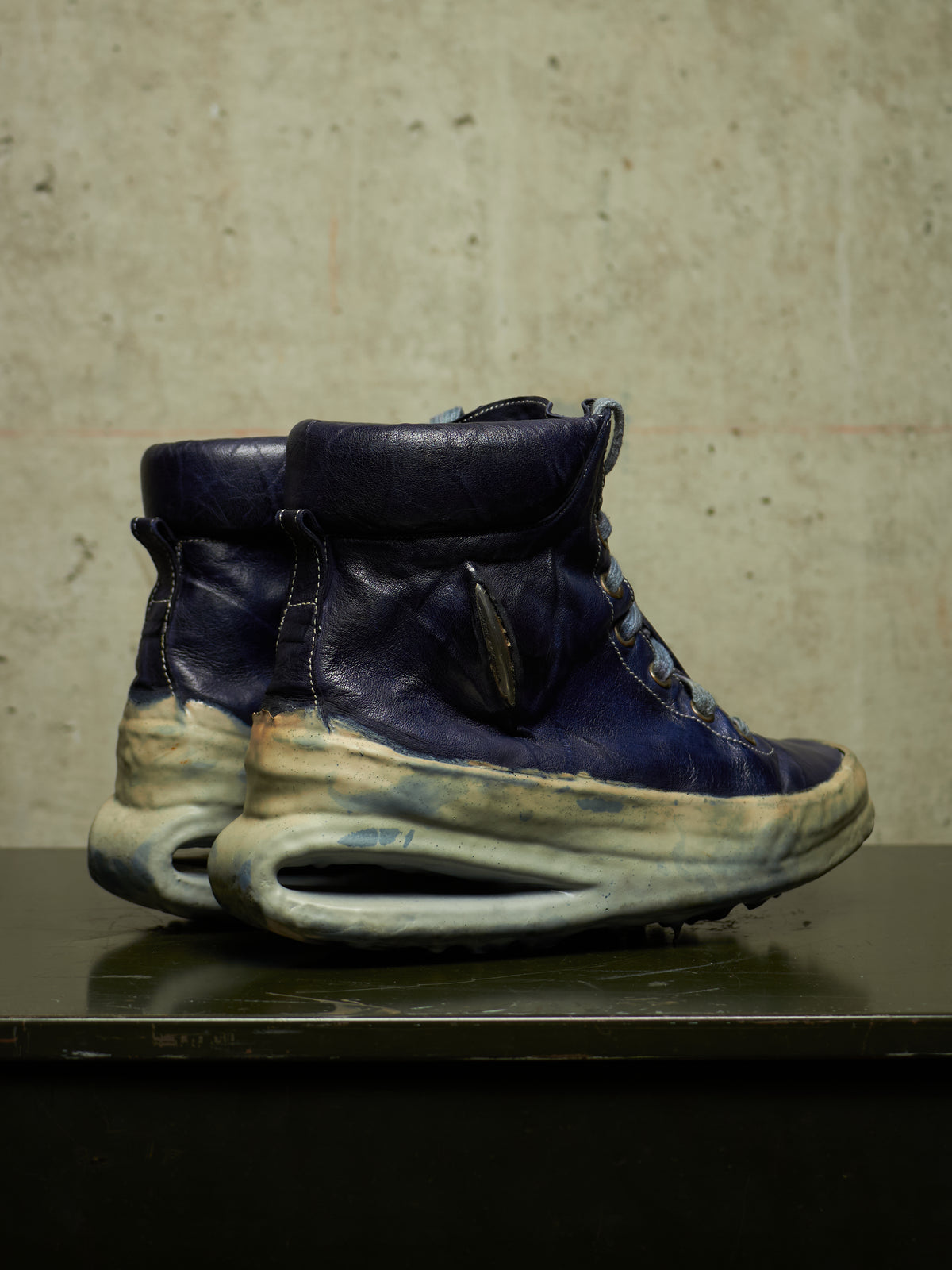A pair of high-top sneakers with dark navy blue leather uppers and thick, sculptural off-white and beige midsoles, displayed on a dark reflective surface against a textured concrete wall.