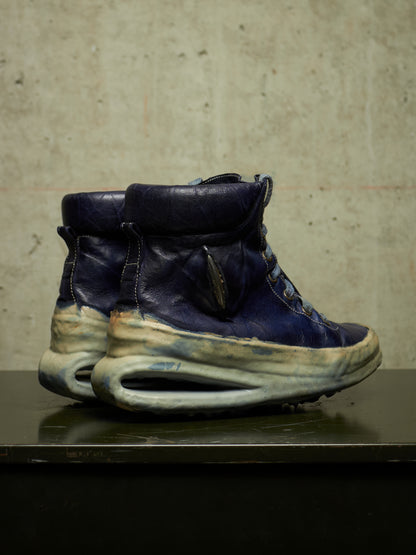 A pair of high-top sneakers with dark navy blue leather uppers and thick, sculptural off-white and beige midsoles, displayed on a dark reflective surface against a textured concrete wall.