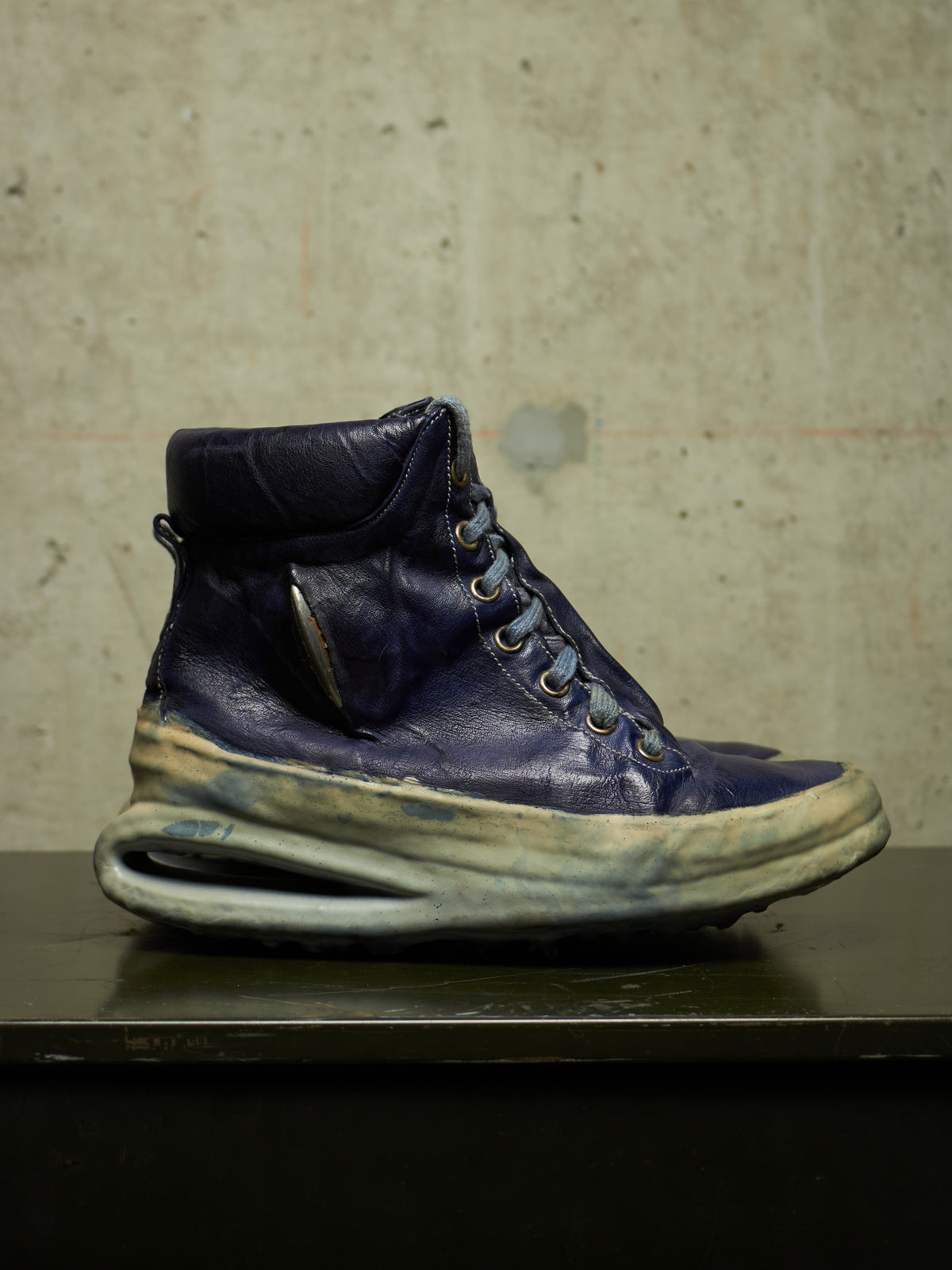 A single high-top sneaker in distressed navy blue leather with a thick, sculpted off-white sole, resting on a dark surface against a textured concrete wall.