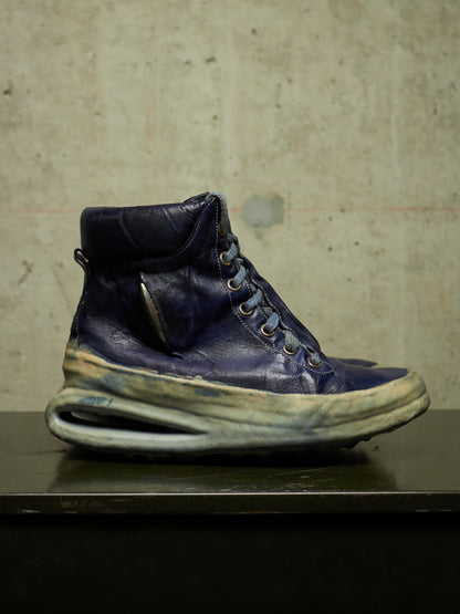 A single high-top sneaker in distressed navy blue leather with a thick, sculpted off-white sole, resting on a dark surface against a textured concrete wall.