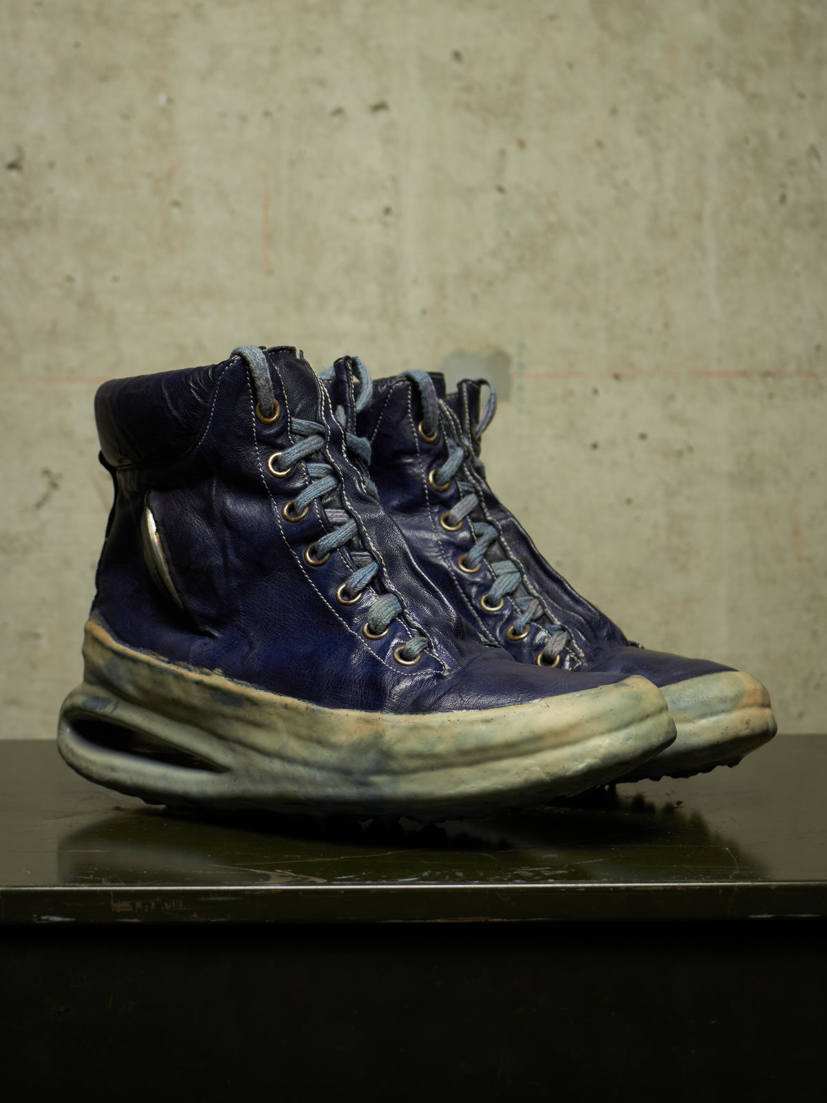 A pair of distressed dark blue leather high-top sneakers with thick, worn off-white midsoles and metal eyelets, displayed on a dark reflective surface against a textured concrete wall.
