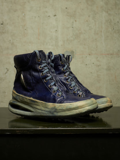 A pair of distressed dark blue leather high-top sneakers with thick, worn off-white midsoles and metal eyelets, displayed on a dark reflective surface against a textured concrete wall.
