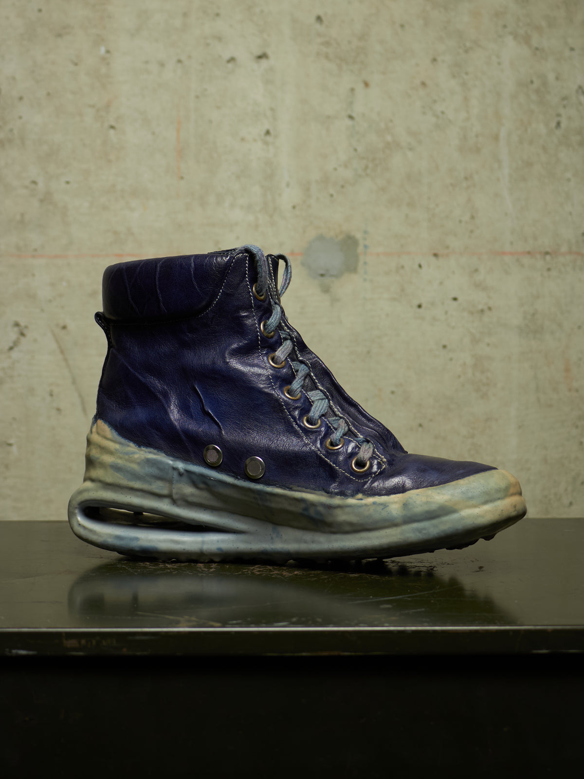 A single high-top sneaker boot in dark navy blue leather with distressed, weathered detailing on the sole and toe, resting on a dark reflective surface against a textured concrete wall.