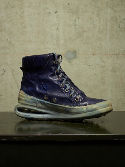 A single high-top sneaker boot in dark navy blue leather with distressed, weathered detailing on the sole and toe, resting on a dark reflective surface against a textured concrete wall.