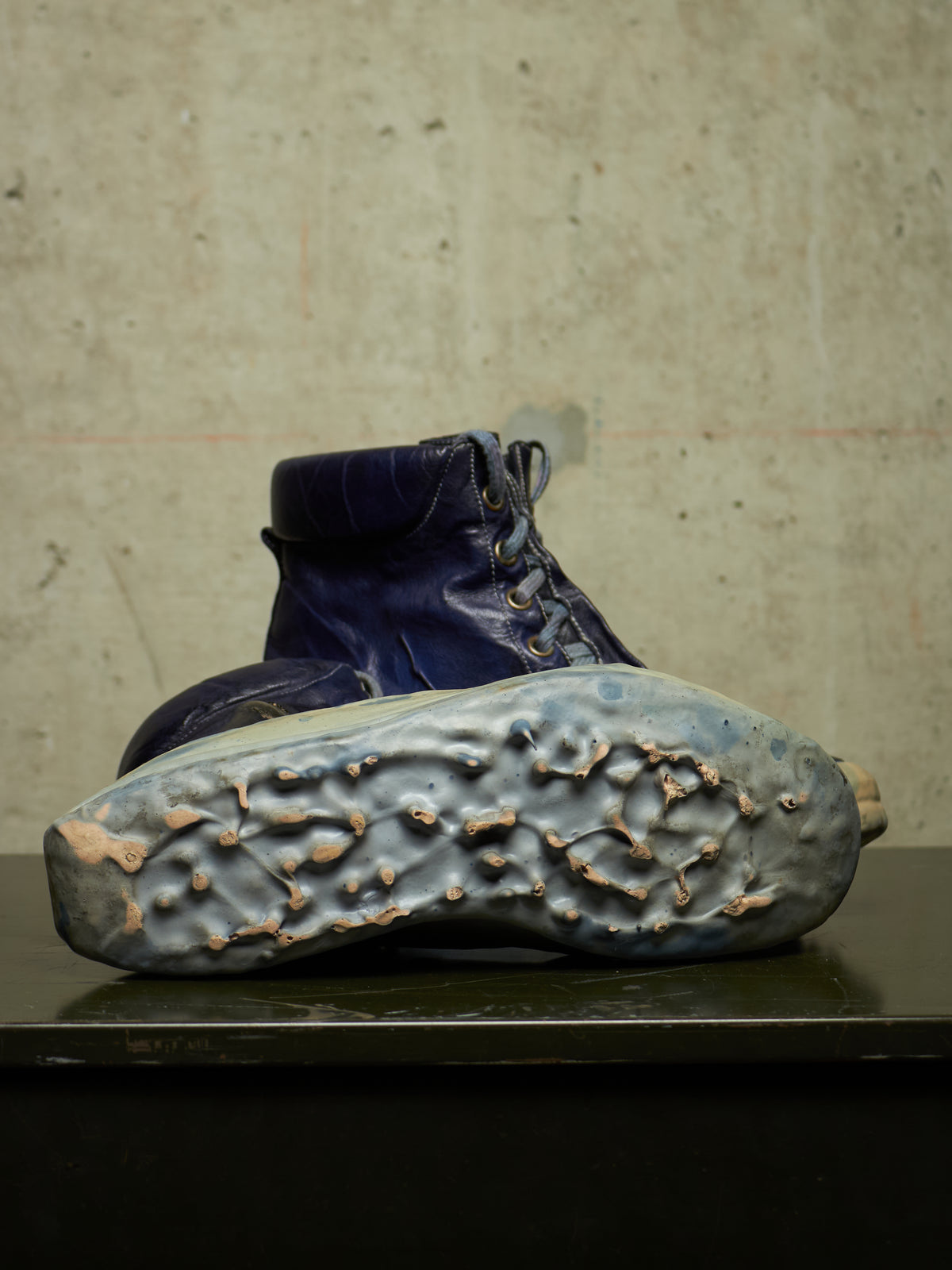 A single dark blue lace-up boot shown from the sole side, resting on a dark surface against a textured concrete wall.